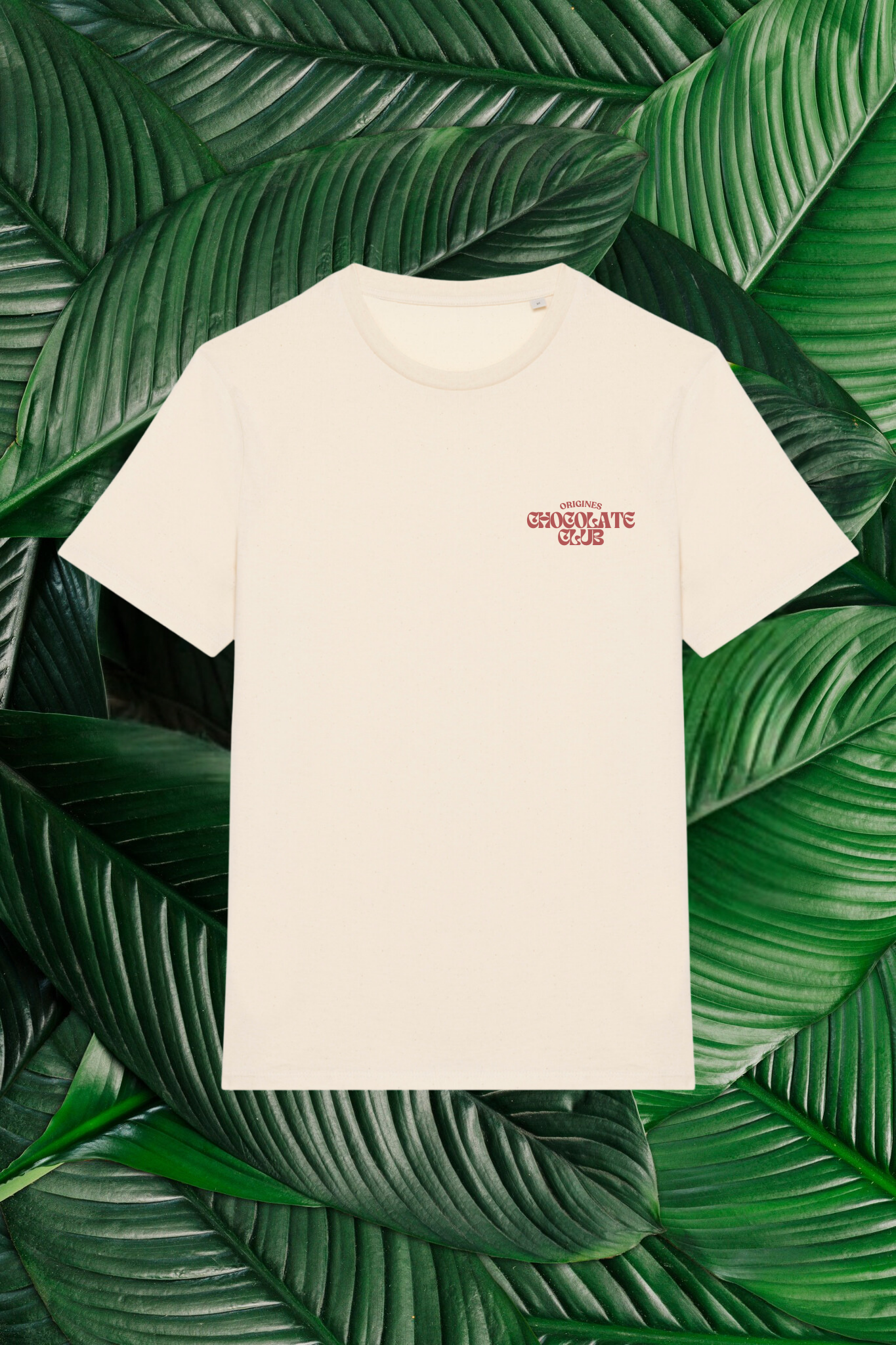 Un t-shirt beige avec un petit logo rouge sur la poitrine, comprenant le texte 'Origines Chocolate Club' dans une police stylisée. Le t-shirt est posé sur un fond composé de grandes feuilles vertes naturelles