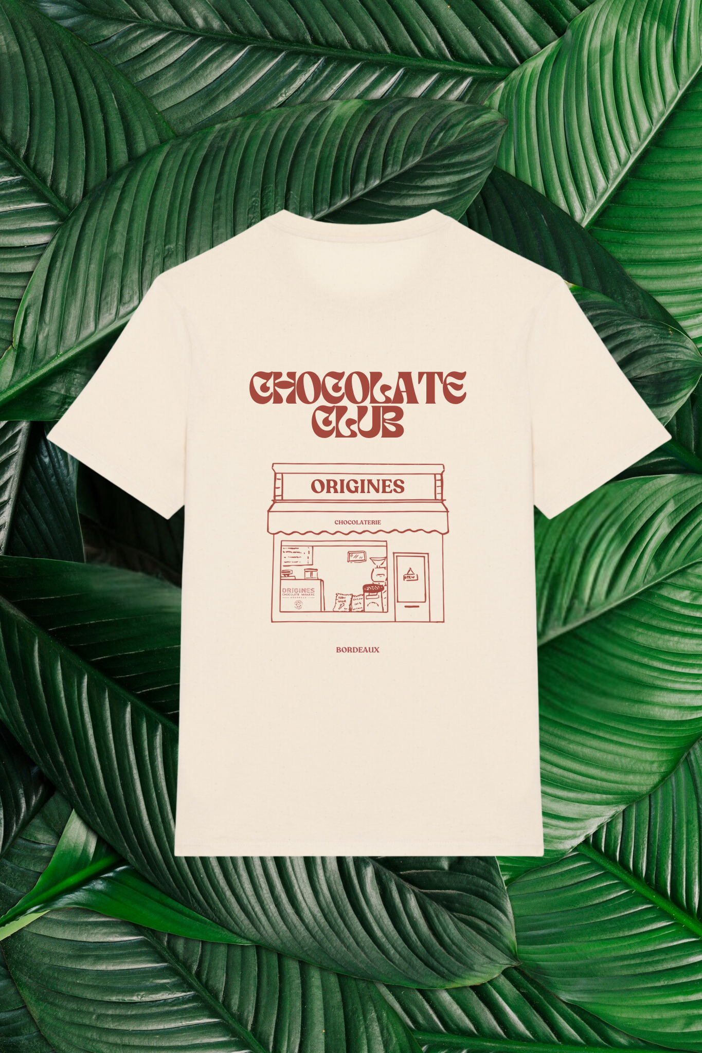 Un t-shirt beige avec un design minimaliste dessiné à la main, représentant une devanture de chocolaterie intitulée 'Origines', accompagné du texte 'Chocolate Club' en lettres stylisées. Le t-shirt est posé sur un fond composé de grandes feuilles vertes naturelles.