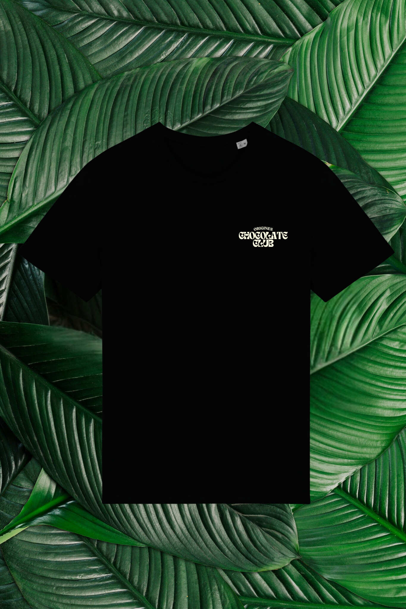 Un t-shirt noir avec un petit logo beige sur la poitrine, comprenant le texte 'Origines Chocolate Club' dans une police stylisée. Le t-shirt est posé sur un fond composé de grandes feuilles vertes naturelles