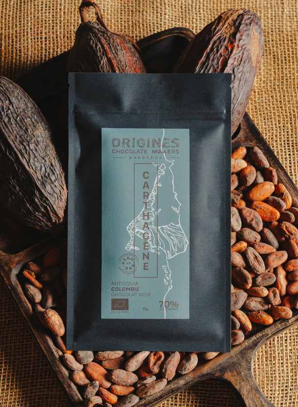 Tablette de chocolat noir Origines Chocolate Makers, emballée dans un sachet noir avec une étiquette bleue indiquant 70 % de cacao provenant de Colombie, entourée de fèves de cacao et de cabosses sur un fond en toile de jute.