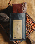 Tablette de chocolat noir Origines Chocolate Makers, emballée dans un sachet noir avec une étiquette jaune indiquant 70 % de cacao provenant du Liberia, entourée de fèves de cacao et de cabosses sur un fond en toile de jute.