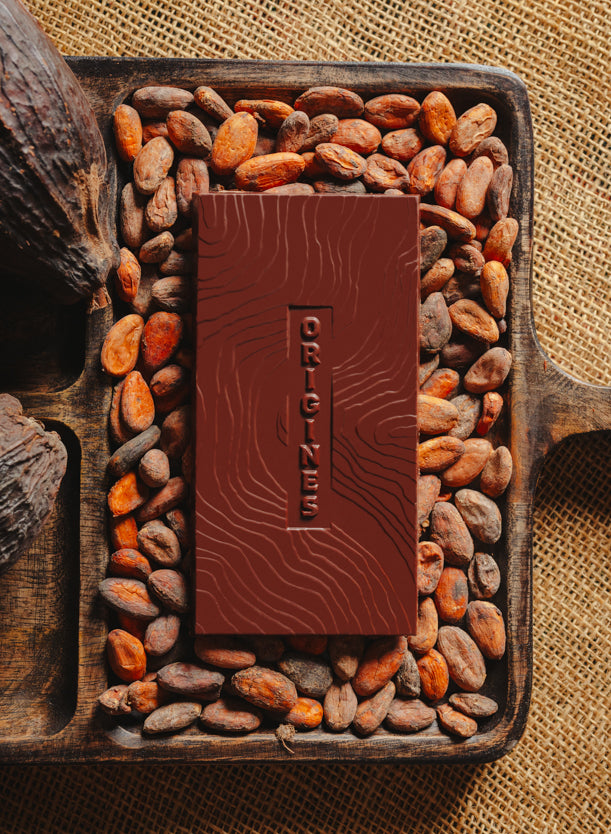 Tablette de chocolat noir Origines Chocolate Makers 70 % de cacao provenant du Pérou, entourée de fèves de cacao et de cabosses sur un fond en toile de jute.