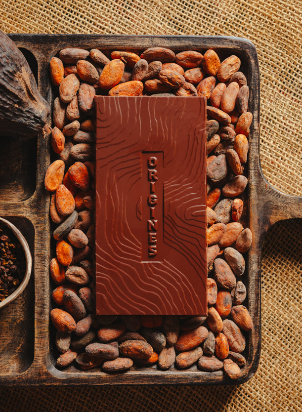 Tablette de chocolat noir au grué  Origines Chocolate Makers 70 % de cacao provenant du Pérou, entourée de fèves de cacao, de cabosses et d'un bol contenant du grué de cacao sur un fond en toile de jute.