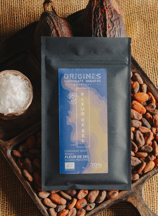 Tablette de chocolat noir à la fleur de sel du bassin d'Arcachon Origines Chocolate Makers, emballée dans un sachet noir avec une étiquette marron indiquant 70 % de cacao provenant du Pérou, entourée de fèves de cacao, de cabosses et d'un bol contenant du sel sur un fond en toile de jute.