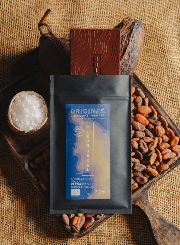 Tablette de chocolat noir à la fleur de sel du bassin d'Arcachon Origines Chocolate Makers, emballée dans un sachet noir avec une étiquette marron indiquant 70 % de cacao provenant du Pérou, entourée de fèves de cacao, de cabosses et d'un bol contenant du sel sur un fond en toile de jute.
