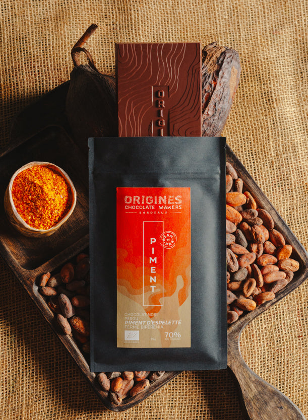 Tablette de chocolat noir au piment d'Espelette Origines Chocolate Makers, emballée dans un sachet noir avec une étiquette orange indiquant 70 % de cacao provenant du Pérou, entourée de fèves de cacao, de cabosses et d'un bol contenant du piment d'Espelette sur un fond en toile de jute.