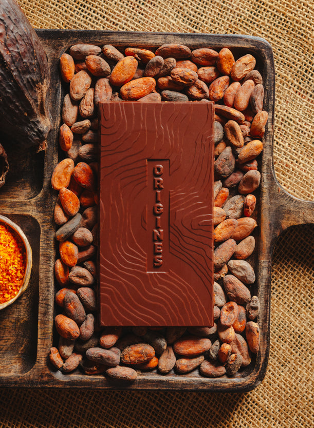 Tablette de chocolat noir au piment d'Espelette Origines Chocolate Makers 70 % de cacao provenant du Pérou, entourée de fèves de cacao, de cabosses et d'un bol contenant du piment d'Espelette sur un fond en toile de jute.