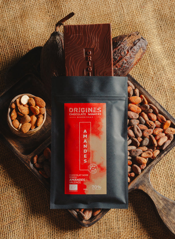 Tablette de chocolat noir au amandes de Valence Origines Chocolate Makers, emballée dans un sachet noir avec une étiquette rouge indiquant 70 % de cacao provenant du Pérou, entourée de fèves de cacao, de cabosses et d'un bol contenant des amandes sur un fond en toile de jute.