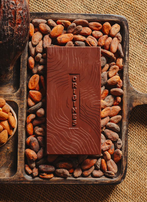 Tablette de chocolat noir au amandes de Valence Origines Chocolate Makers 70 % de cacao provenant du Pérou, entourée de fèves de cacao, de cabosses et d'un bol contenant des amandes sur un fond en toile de jute.