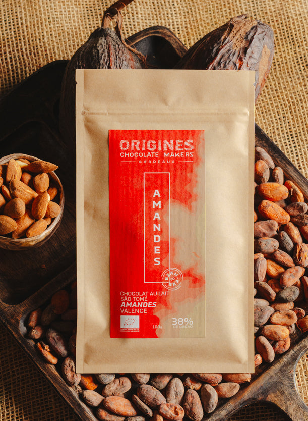 Tablette de chocolat au lait au amandes de Valence Origines Chocolate Makers, emballée dans un sachet kraft avec une étiquette rouge indiquant 38 % de cacao de São Tomé entourée de fèves de cacao, de cabosses et d'un bol contenant des amandes sur un fond en toile de jute.