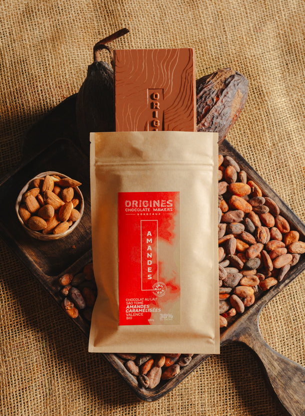 Tablette de chocolat au lait au amandes de Valence Origines Chocolate Makers, emballée dans un sachet kraft avec une étiquette rouge indiquant 38 % de cacao de São Tomé entourée de fèves de cacao, de cabosses et d'un bol contenant des amandes sur un fond en toile de jute.