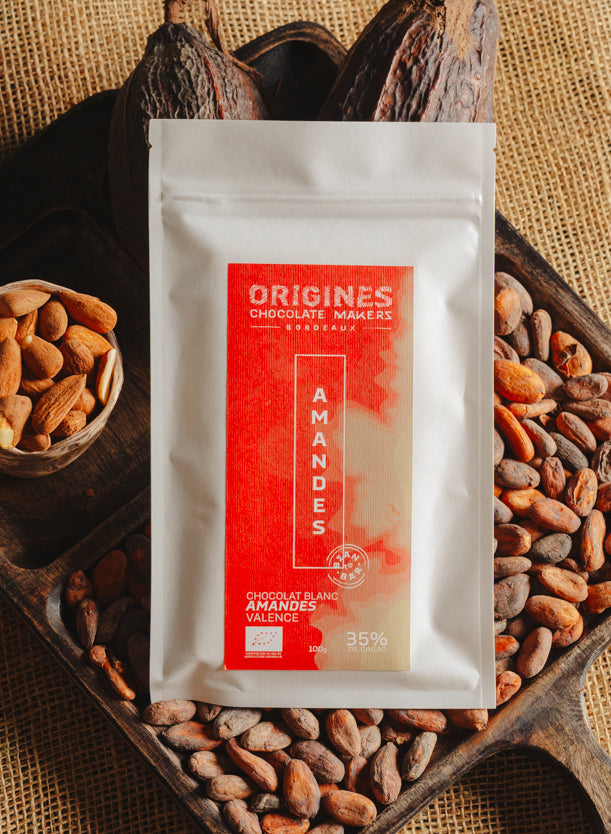 Tablette de chocolat blanc aux amandes de Valence Origines Chocolate Makers, emballée dans un sachet blanc avec une étiquette marron indiquant 35 % de cacao provenant du Pérou, entourée de fèves de cacao, de cabosses et d'un bol contenant des amandes sur un fond en toile de jute.