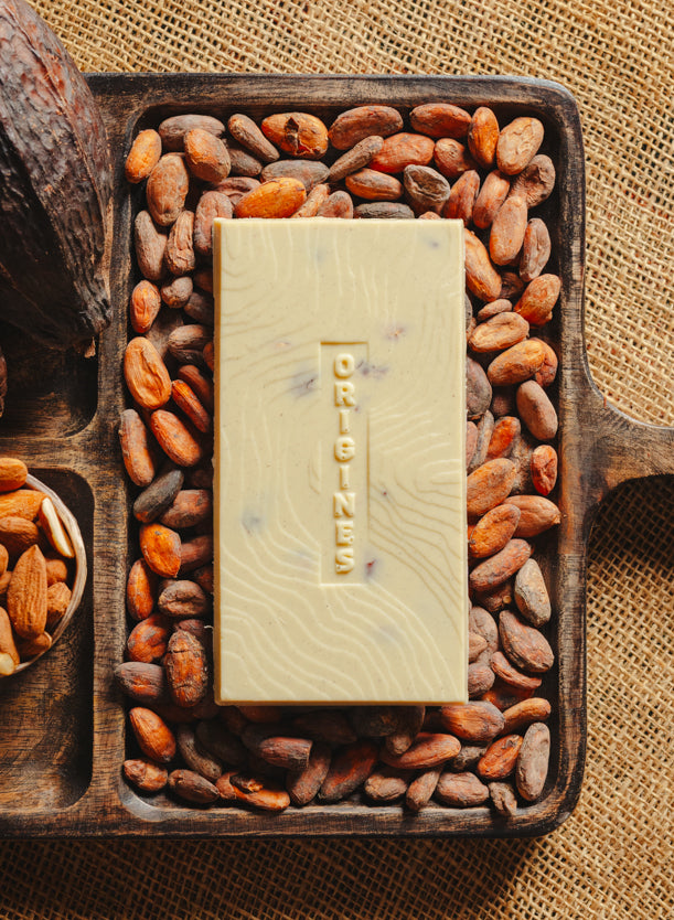Tablette de chocolat blanc aux amandes de Valence Origines Chocolate Makers 35 % de cacao provenant du Pérou, entourée de fèves de cacao, de cabosses et d'un bol contenant des amandes sur un fond en toile de jute.