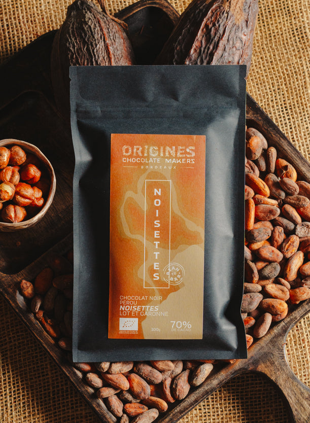 Tablette de chocolat noir au noisettes de lot et garonne Origines Chocolate Makers, emballée dans un sachet noir avec une étiquette marron indiquant 70 % de cacao provenant du Pérou, entourée de fèves de cacao, de cabosses et d'un bol contenant des noisettes sur un fond en toile de jute.