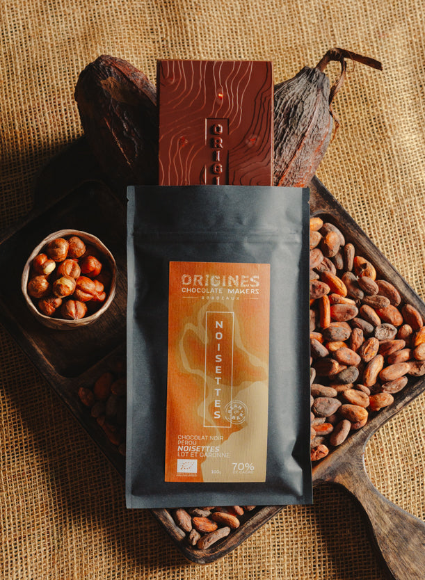 Tablette de chocolat noir au noisettes de lot et garonne Origines Chocolate Makers, emballée dans un sachet noir avec une étiquette marron indiquant 70 % de cacao provenant du Pérou, entourée de fèves de cacao, de cabosses et d'un bol contenant des noisettes sur un fond en toile de jute.
