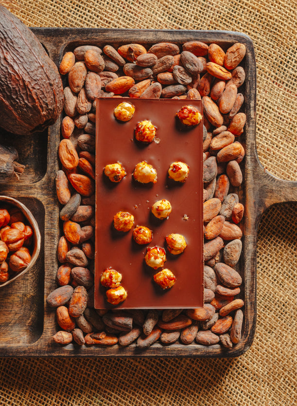 Dos d'une tablette de chocolat noir avec des noisettes de lot et garonne Origines Chocolate Makers 70 % de cacao provenant du Pérou, entourée de fèves de cacao, de cabosses et d'un bol contenant des noisettes sur un fond en toile de jute.