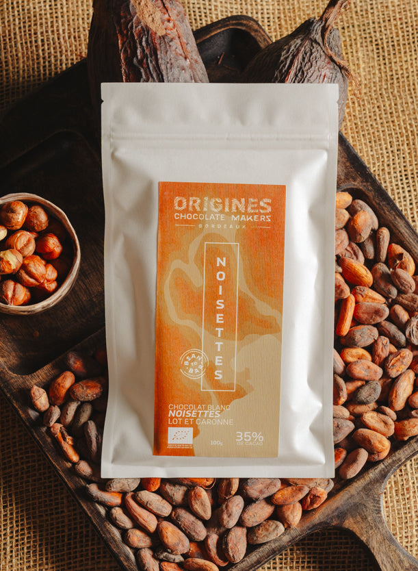 Tablette de chocolat blanc au noisettes de lot et garonne Origines Chocolate Makers, emballée dans un sachet blanc avec une étiquette marron indiquant 35 % de cacao, entourée de fèves de cacao, de cabosses et d'un bol contenant des noisettes sur un fond en toile de jute.