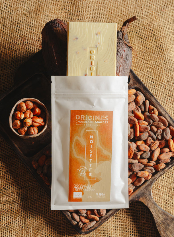 Tablette de chocolat blanc au noisettes de lot et garonne Origines Chocolate Makers, emballée dans un sachet blanc avec une étiquette marron indiquant 35 % de cacao entourée de fèves de cacao, de cabosses et d'un bol contenant des noisettes sur un fond en toile de jute.