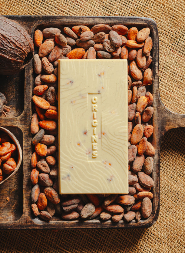 Tablette de chocolat blanc au noisettes de lot et garonne Origines Chocolate Makers 35 % de cacao, entourée de fèves de cacao, de cabosses et d'un bol contenant des noisettes sur un fond en toile de jute.