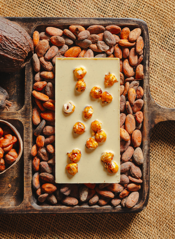 Dos d'une tablette de chocolat blanc au noisettes de lot et garonne Origines Chocolate Makers 35 % de cacao, entourée de fèves de cacao, de cabosses et d'un bol contenant des noisettes sur un fond en toile de jute.