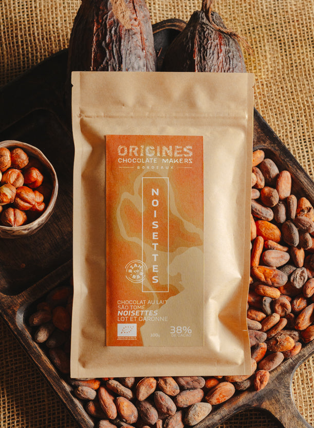 Tablette de chocolat au lait au noisettes de lot et garonne Origines Chocolate Makers, emballée dans un sachet kraft avec une étiquette marron indiquant 38 % de cacao de São Tomé entourée de fèves de cacao, de cabosses et d'un bol contenant des noisettes sur un fond en toile de jute.
