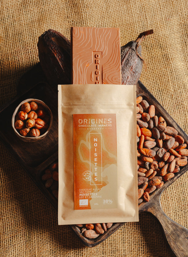 Tablette de chocolat au lait au noisettes de lot et garonne Origines Chocolate Makers, emballée dans un sachet kraft avec une étiquette marron indiquant 38 % de cacao de Sâo Tomé entourée de fèves de cacao, de cabosses et d'un bol contenant des noisettes sur un fond en toile de jute.