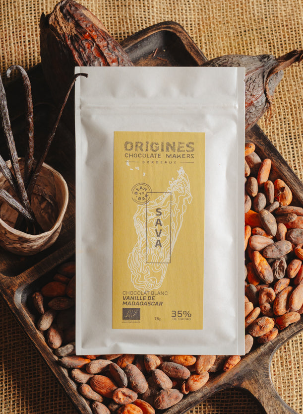 Tablette de chocolat blanc à la vanille Origines Chocolate Makers, emballée dans un sachet blanc avec une étiquette jaune indiquant 35% de cacao provenant de Madagascar, entourée de fèves de cacao, de gousses de vanille et de cabosses sur un fond en toile de jute.