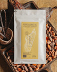 Tablette de chocolat blanc à la vanille Origines Chocolate Makers, emballée dans un sachet blanc avec une étiquette jaune indiquant 35% de cacao provenant de Madagascar, entourée de fèves de cacao, de gousses de vanille et de cabosses sur un fond en toile de jute.