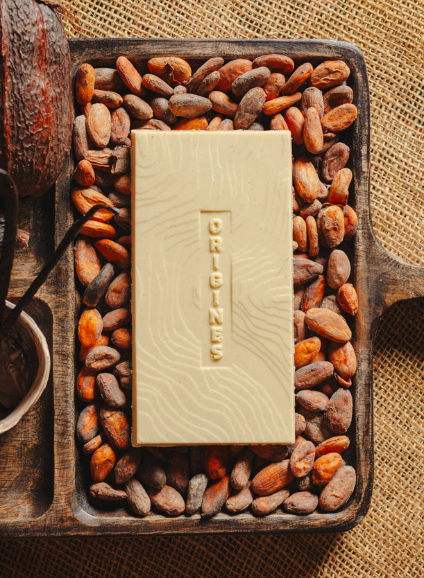 Tablette de chocolat blanc à la vanille Origines Chocolate Makers 35% de cacao provenant de Madagascar, entourée de fèves de cacao, de gousses de vanille et de cabosses sur un fond en toile de jute.