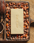 Tablette de chocolat blanc à la vanille Origines Chocolate Makers 35% de cacao provenant de Madagascar, entourée de fèves de cacao, de gousses de vanille et de cabosses sur un fond en toile de jute.
