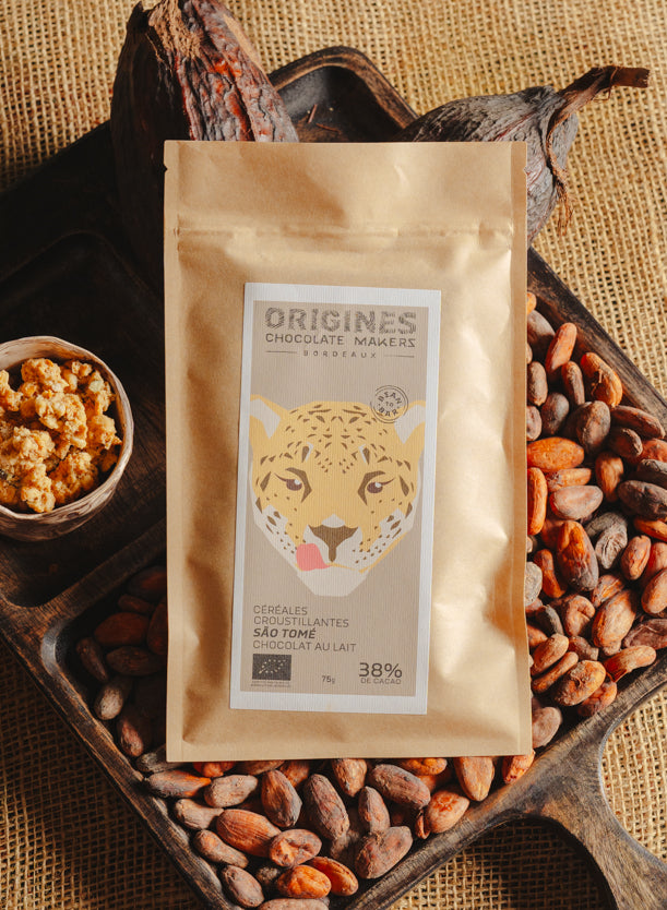 Tablette de chocolat au lait au céréales croustillantes Origines Chocolate Makers, emballée dans un sachet kraft avec une étiquette représentant un tigre indiquant 38 % de cacao provenant de Sao Tome, entourée de fèves de cacao, de cabosses et d'un bol contenant des céréales sur un fond en toile de jute.