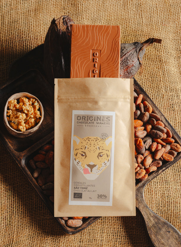 Tablette de chocolat au lait au céréales croustillantes Origines Chocolate Makers, emballée dans un sachet kraft avec une étiquette représentant un tigre indiquant 38 % de cacao provenant de Sao Tome, entourée de fèves de cacao, de cabosses et d'un bol contenant des céréales sur un fond en toile de jute.