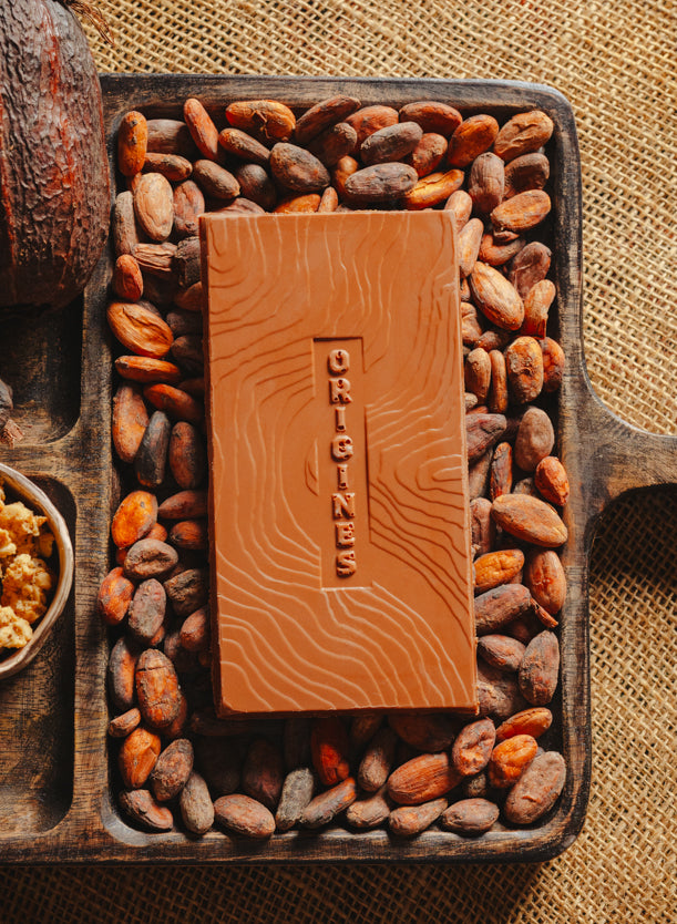 Tablette de chocolat au lait au céréales croustillantes Origines Chocolate Makers, 38 % de cacao provenant de Sao Tome, entourée de fèves de cacao, de cabosses et d'un bol contenant des céréales sur un fond en toile de jute.