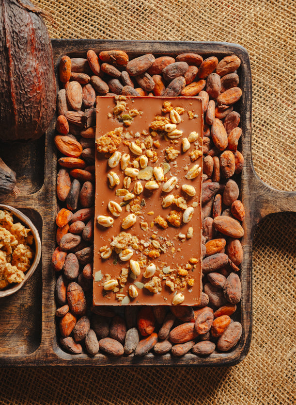 Dos d'une tablette de chocolat au lait au céréales croustillantes Origines Chocolate Makers, 38 % de cacao provenant de Sao Tome, entourée de fèves de cacao, de cabosses et d'un bol contenant des céréales sur un fond en toile de jute.