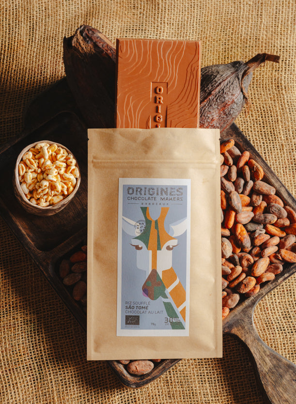 Tablette de chocolat au lait au riz soufflé Origines Chocolate Makers, emballée dans un sachet kraft avec une étiquette représentant une girafe indiquant 38 % de cacao provenant de Sao Tome, entourée de fèves de cacao, de cabosses et d'un bol contenant du riz soufflé sur un fond en toile de jute.