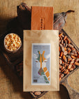 Tablette de chocolat au lait au riz soufflé Origines Chocolate Makers, emballée dans un sachet kraft avec une étiquette représentant une girafe indiquant 38 % de cacao provenant de Sao Tome, entourée de fèves de cacao, de cabosses et d'un bol contenant du riz soufflé sur un fond en toile de jute.