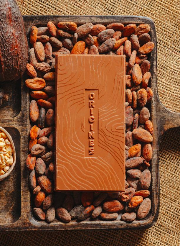 Tablette de chocolat au lait au riz soufflé Origines Chocolate Makers,  38 % de cacao provenant de Sao Tome, entourée de fèves de cacao, de cabosses et d'un bol contenant du riz soufflé sur un fond en toile de jute.