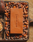 Tablette de chocolat au lait au riz soufflé Origines Chocolate Makers,  38 % de cacao provenant de Sao Tome, entourée de fèves de cacao, de cabosses et d'un bol contenant du riz soufflé sur un fond en toile de jute.