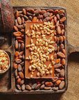 Dos d'une tablette de chocolat au lait au riz soufflé Origines Chocolate Makers 38 % de cacao provenant de Sao Tome, entourée de fèves de cacao, de cabosses et d'un bol contenant du riz soufflé sur un fond en toile de jute.