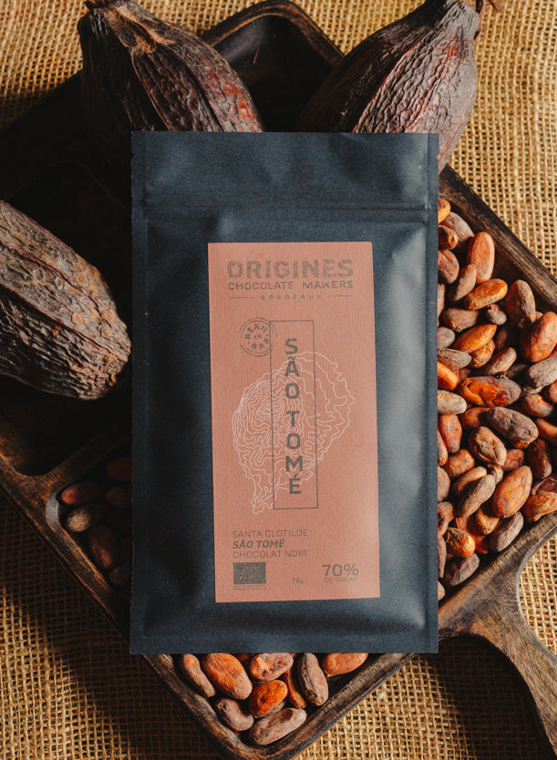 Tablette de chocolat noir Origines Chocolate Makers, emballée dans un sachet noir avec une étiquette rouge indiquant 70 % de cacao provenant de Sao Tome, entourée de fèves de cacao et de cabosses sur un fond en toile de jute.