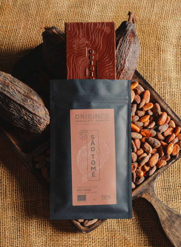 Tablette de chocolat noir Origines Chocolate Makers, emballée dans un sachet noir avec une étiquette rouge indiquant 70 % de cacao provenant de Sao Tome, entourée de fèves de cacao et de cabosses sur un fond en toile de jute.