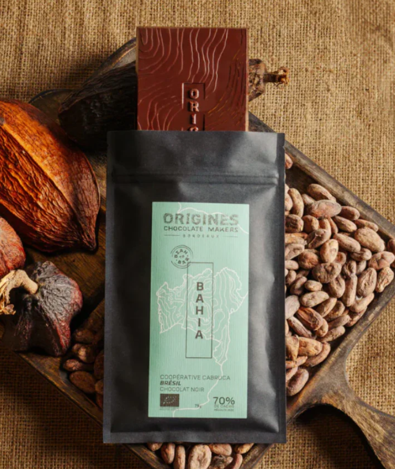 Tablette de chocolat noir Origines Chocolate Makers, emballée dans un sachet noir avec une étiquette bleue indiquant 70 % de cacao provenant de la coopérative Cabruca à Bahia, Brésil, entourée de fèves de cacao et de cabosses sur un fond en toile de jute.