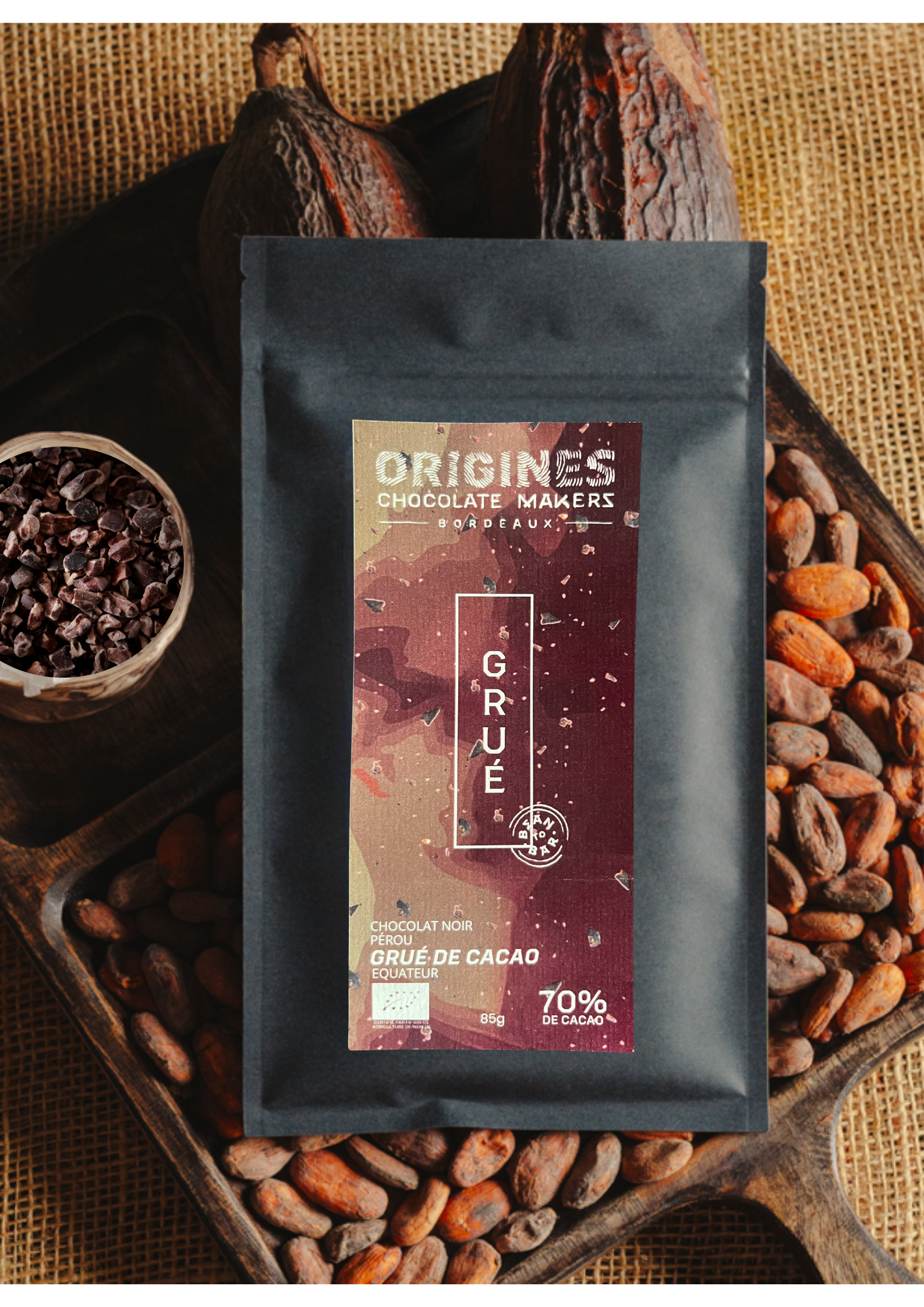 Tablette de chocolat noir au grué  Origines Chocolate Makers, emballée dans un sachet noir avec une étiquette rouge indiquant 70 % de cacao provenant du Pérou, entourée de fèves de cacao, de cabosses et d'un bol contenant du grué de cacao sur un fond en toile de jute.