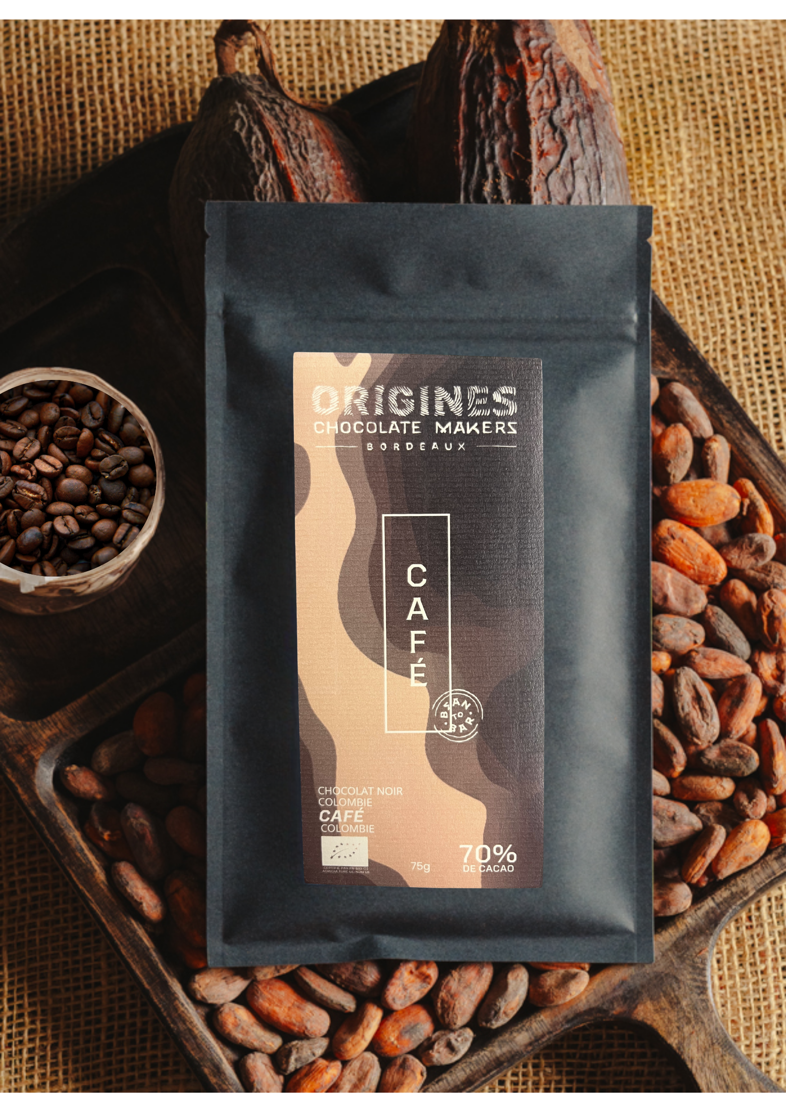 Tablette de chocolat noir au café  Origines Chocolate Makers, emballée dans un sachet noir avec une étiquette marron indiquant 70 % de cacao provenant de Colombie, entourée de fèves de cacao, de cabosses et d'un bol contenant du café sur un fond en toile de jute.