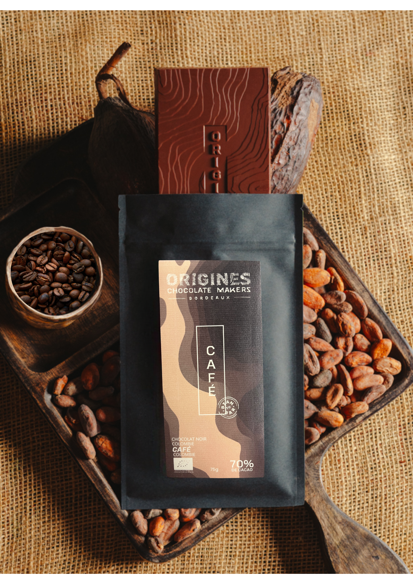 Tablette de chocolat noir au café  Origines Chocolate Makers, emballée dans un sachet noir avec une étiquette marron indiquant 70 % de cacao provenant de Colombie, entourée de fèves de cacao, de cabosses et d'un bol contenant du café sur un fond en toile de jute.