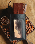 Tablette de chocolat noir au café  Origines Chocolate Makers, emballée dans un sachet noir avec une étiquette marron indiquant 70 % de cacao provenant de Colombie, entourée de fèves de cacao, de cabosses et d'un bol contenant du café sur un fond en toile de jute.