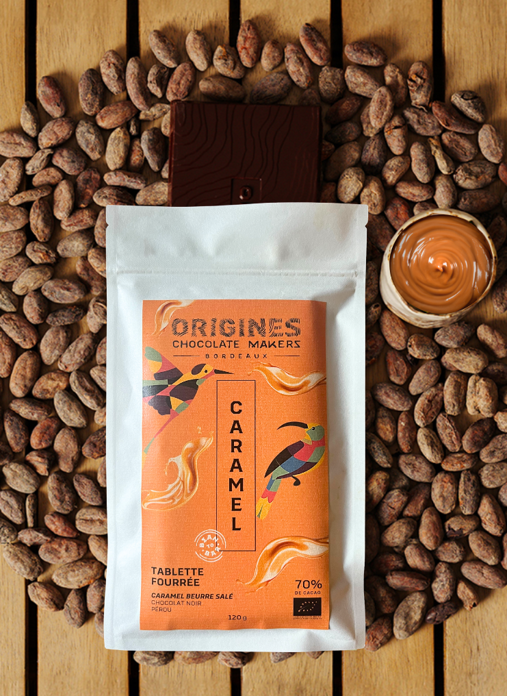 Tablette de chocolat noir Origines Chocolate Makers au caramel, emballée dans un sachet orange orné de toucans colorés, entourée de fèves de cacao et d'un petit bol de caramel sur une surface en bois.