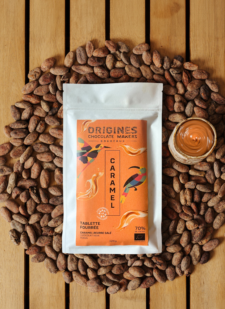 Tablette de chocolat noir Origines Chocolate Makers au caramel, emballée dans un sachet orange orné de toucans colorés, entourée de fèves de cacao et d'un petit bol de caramel sur une surface en bois.