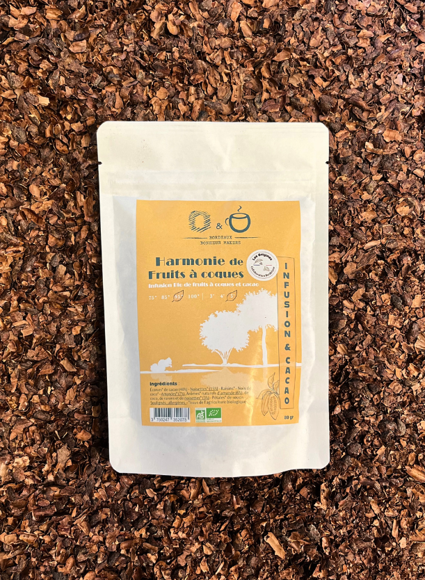 Un sachet d'infusion au cacao cacao en papier blanc avec une étiquette jaune indiquant Harmonie de Fruits à Coques avec l'inscription "Origines Chocolate Maker Bordeaux" posé dans du grué de cacao. 