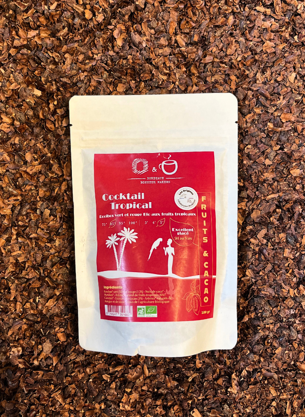 Un sachet d'infusion au cacao cacao en papier blanc avec une étiquette rouge indiquant Cocktail Tropical avec l'inscription "Origines Chocolate Maker Bordeaux" posé dans du grué de cacao. 